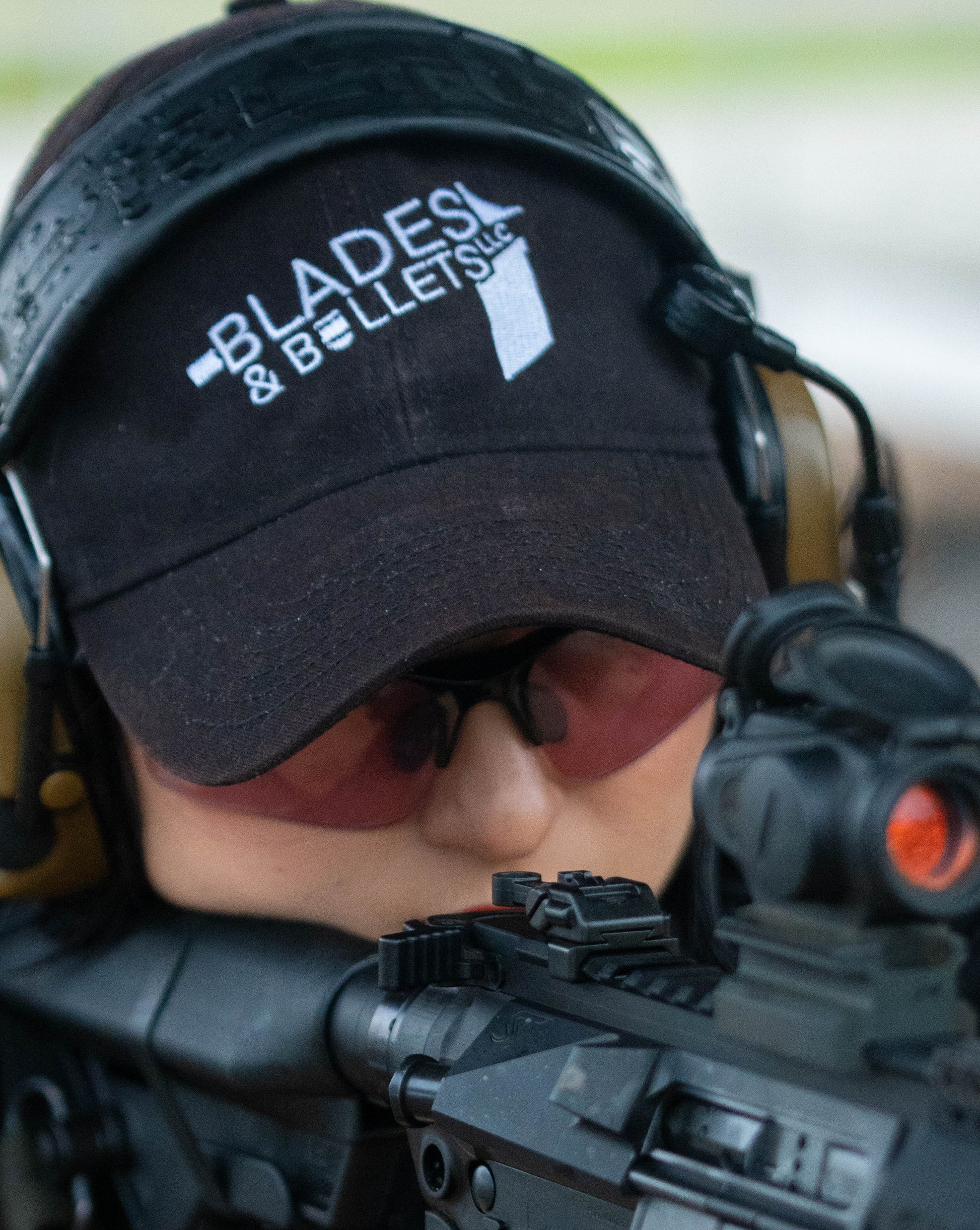 A photo of Sierrah with a rifle, promoting the Blades and Bullets Hat for sale in the online shop.
